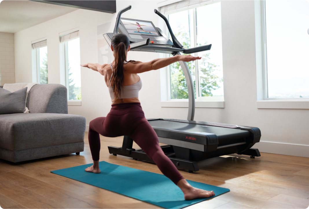 woman doing yoga next to her nordictrack incline trainer while an ifit trainer is displayed on the hd touchscreen